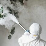 Person in protective gear fumigating an indoor plant, emitting smoke.